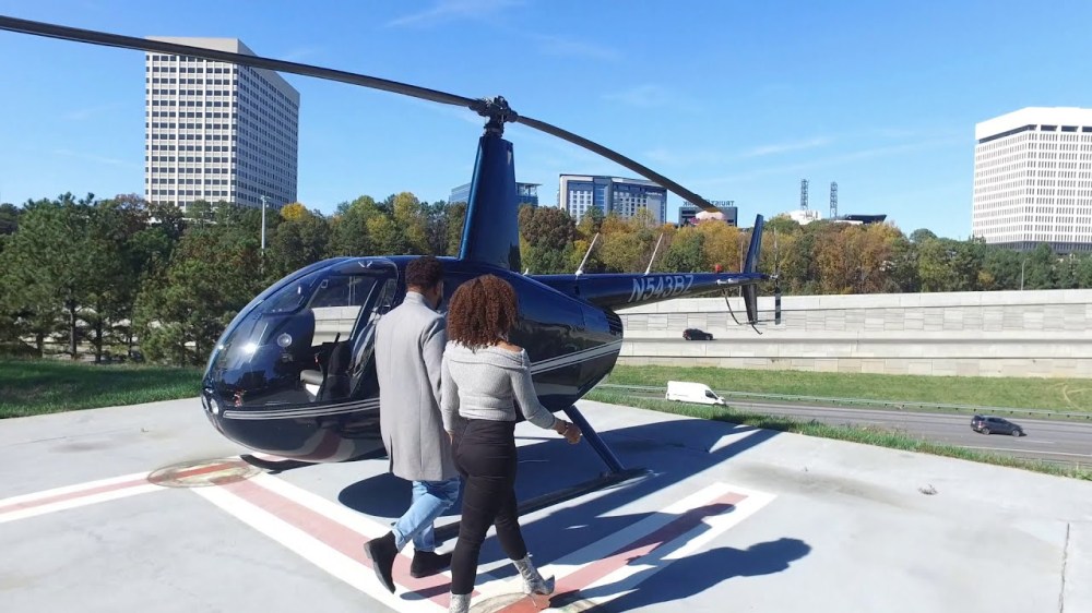 Couple boarding a parked helicopter on a helipad with buildings and trees in the background.