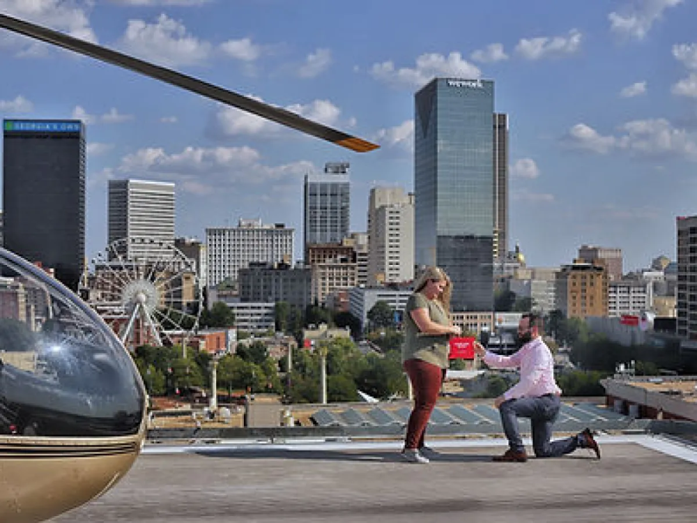 Man kneeling and proposing to woman on rooftop with city skyline and helicopter.